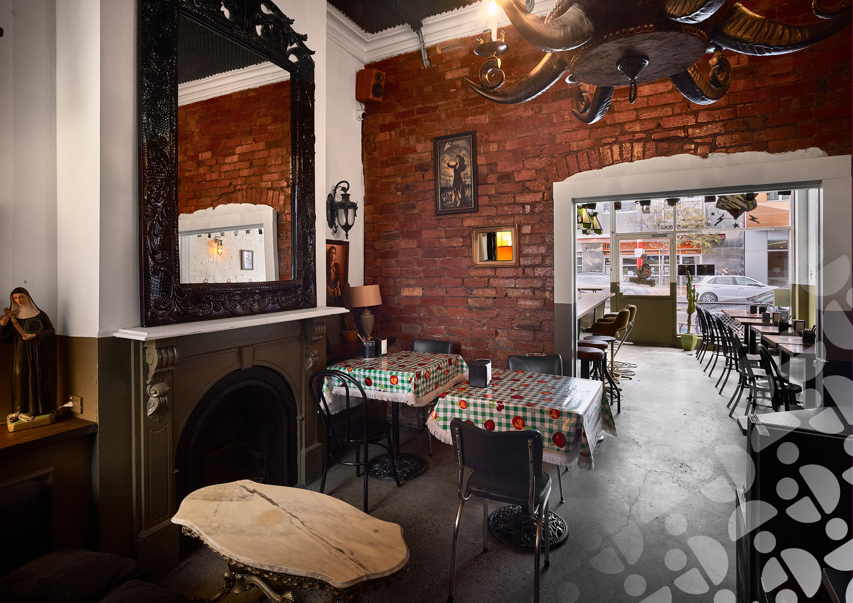 Cozy cafe interior with exposed brick wall, ornate black-framed mirror, and brick fireplace; tables covered in red-green checkered cloths and black chairs nearby, view to street through doorway.