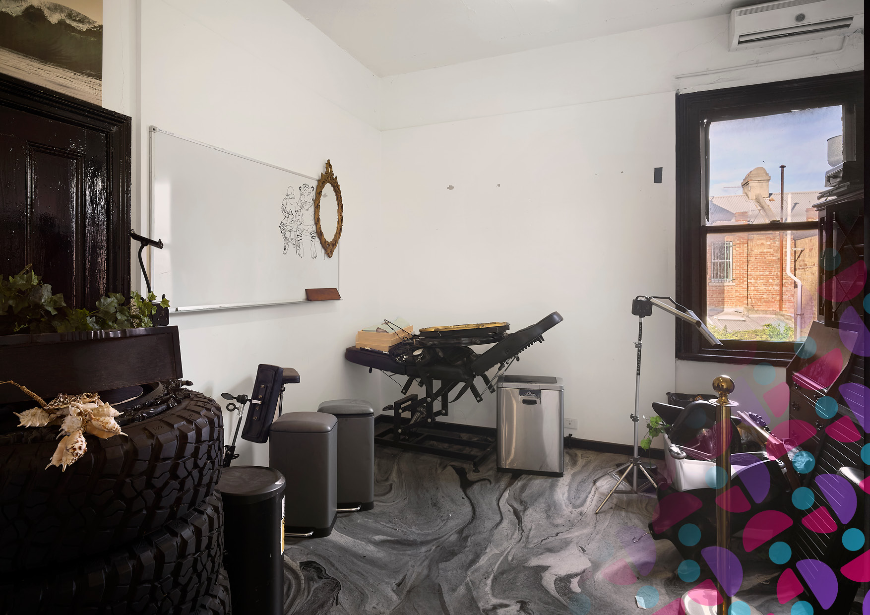 Bright studio with a workout bench and assorted equipment, a whiteboard with a sketch, and an ornate mirror on white walls near a large window showing brick buildings outside.