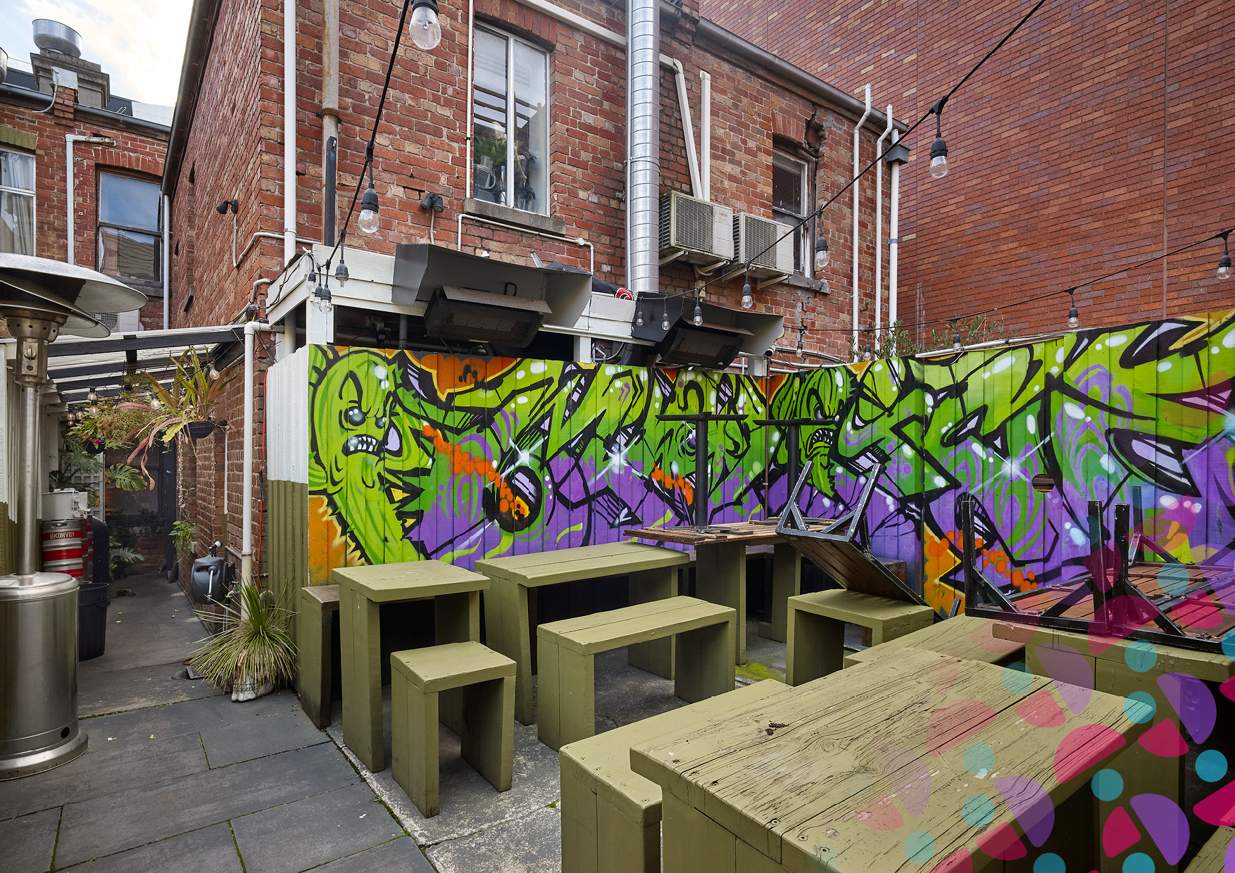 Outdoor courtyard with lime-green benches and wooden tables beneath string lights, in front of a colorful graffiti wall on brick buildings.