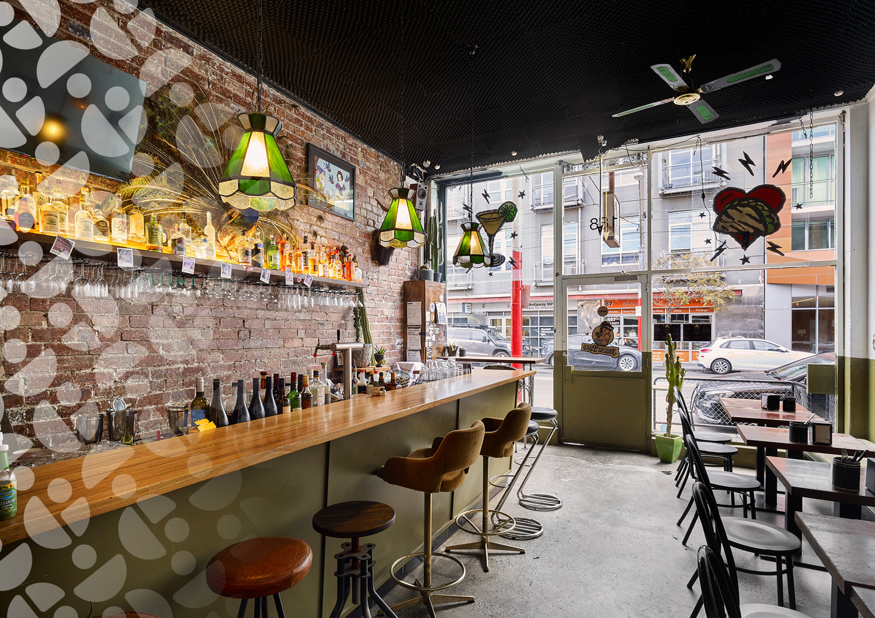 Brick-walled bar interior with liquor on a shelf, green pendant lights, and a long wooden counter with stools.