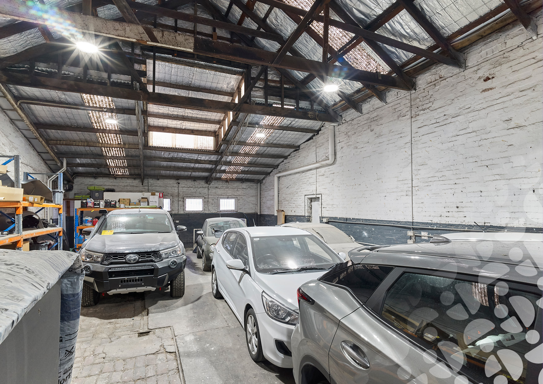 Interior of a workshop/garage with parked cars, white brick walls, and exposed wooden beams overhead.