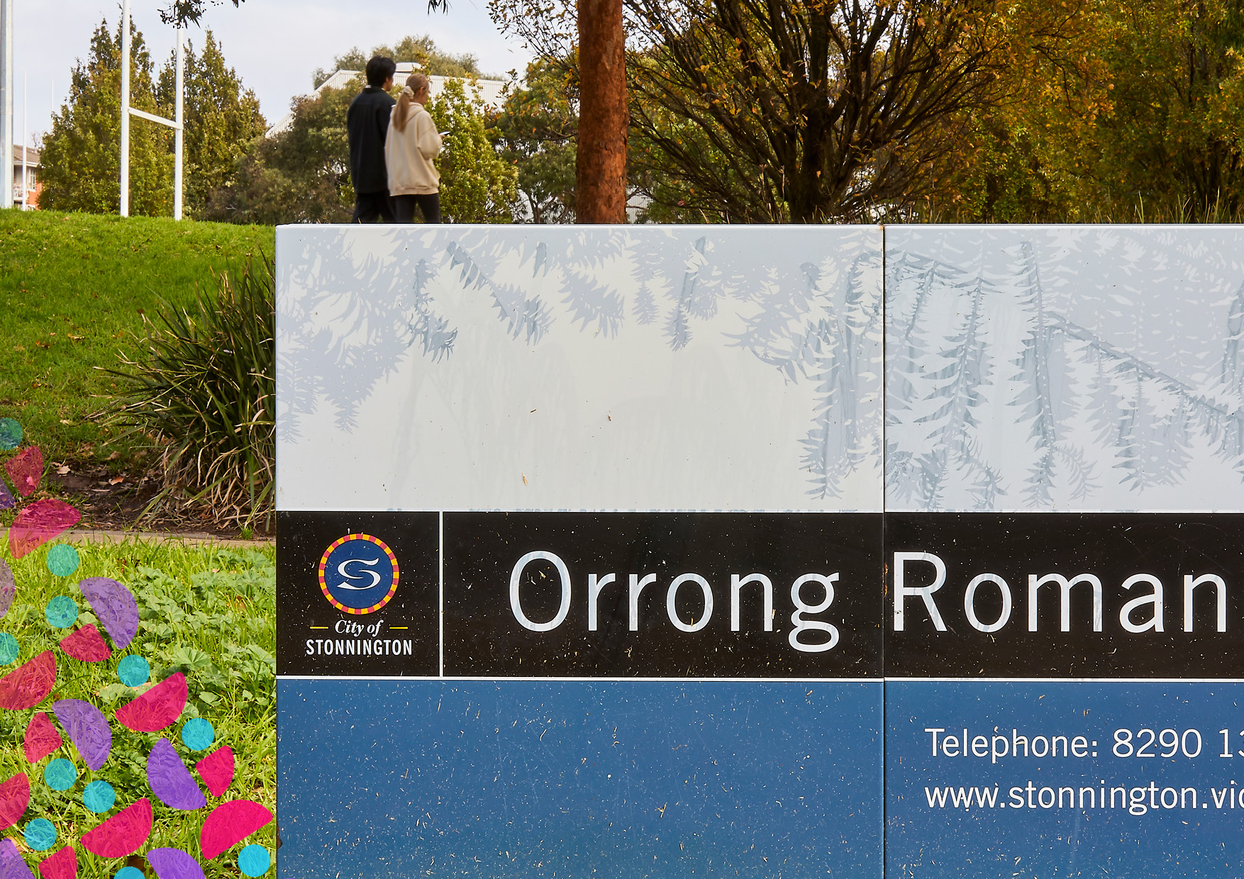 Park sign with fern artwork and the words 'Orrong Roman' and 'City of Stonnington'; two people walk in the background on grass.